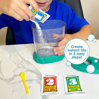 A child wearing a blue shirt is holding the aquarium with one hand, and pouring powder from a small packed labled with the number 1 with the other hand. The lid of the aquarium, a packet with the number 2, a packet with the number 3, and a yellow plastic spoon all lay on the surface in front of the child. There is a callout in a circle that reads "Create instant life in 3 easy steps!"
