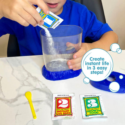 A child wearing a blue shirt is holding the Magiquarium with one hand, and pouring powder from a small packet labled with the number 1 with the other hand. The lid of the aquarium, a packet with the number 2, a packet with the number 3, and a yellow plastic spoon all lay on the surface in front of the child.