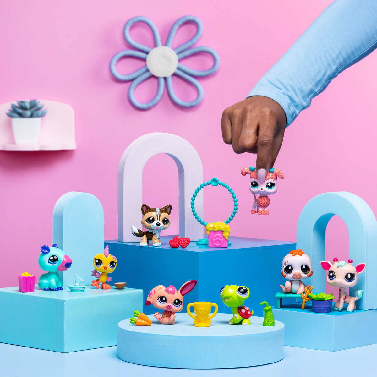 A playful scene featuring several 'Littlest Pet Shop' toy figures arranged on pastel blue and pink geometric platforms against a pink background. A hand is holding one of the pet figures, a pink dog, in mid-air. Other toys include a blue and pink creature with a yellow drink, a yellow bird with a blue cup, a brown dog with red accessories, a green turtle with a trophy, a pink bunny with a carrot, a white and orange puppy, and a white and pink deer-like figure. The background is decorated with a 