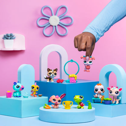 A playful scene featuring several 'Littlest Pet Shop' toy figures arranged on pastel blue and pink geometric platforms against a pink background. A hand is holding one of the pet figures, a pink dog, in mid-air. Other toys include a blue and pink creature with a yellow drink, a yellow bird with a blue cup, a brown dog with red accessories, a green turtle with a trophy, a pink bunny with a carrot, a white and orange puppy, and a white and pink deer-like figure. The background is decorated with a 